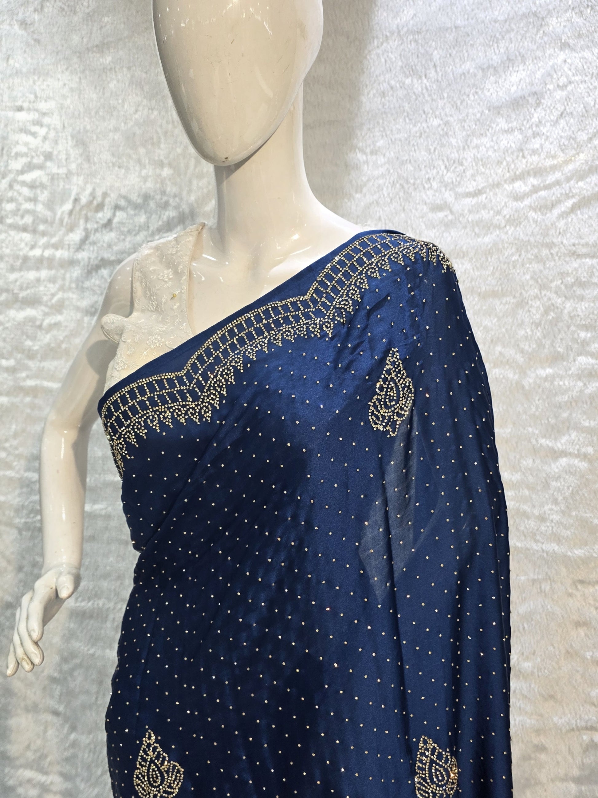 A stunning royal blue pure satin silk saree featuring rich allover stone work buttis that create a glamorous shimmer across the body. The highlight of this saree is its heavy stone-work necklace border, adding a premium designer touch. This saree comes with a matching self royal blue blouse, making it an ideal pick for evening events, parties, and wedding ceremonies where bold elegance stands out.